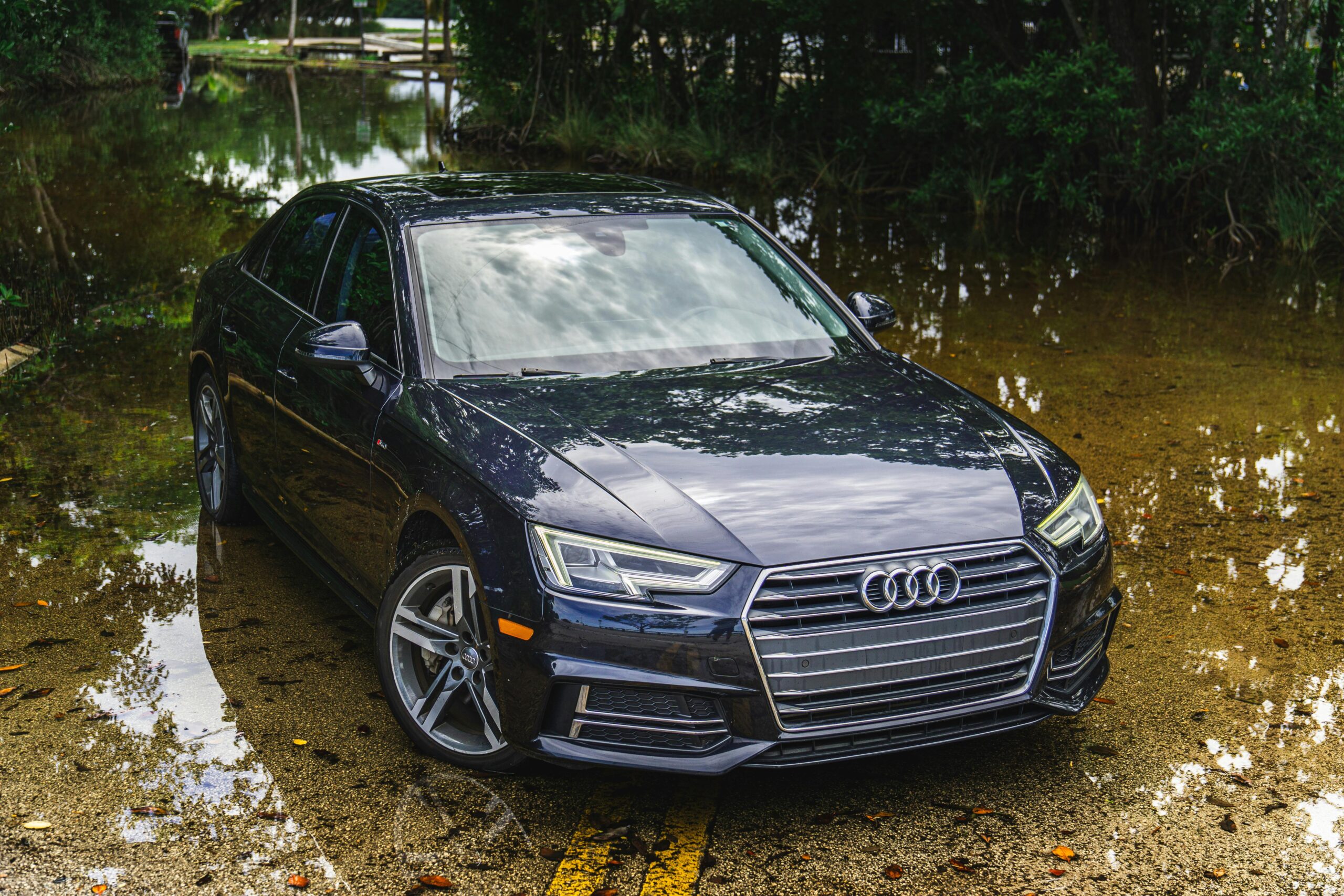 A black sedan car parked on a flooded road surrounded by reflections and trees.